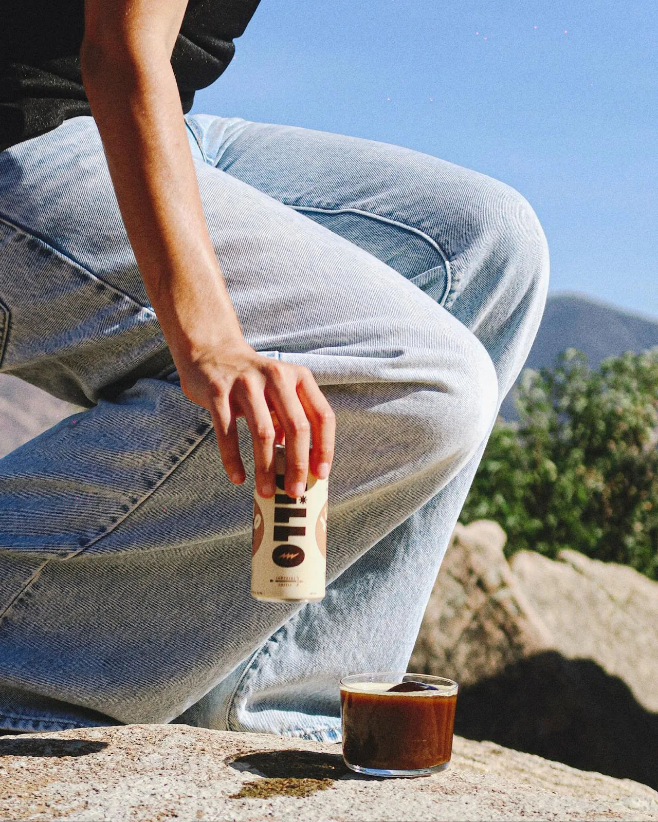 A person in light blue jeans holding a JiLLO can beside an iced coffee cocktail on sunlit rocks
