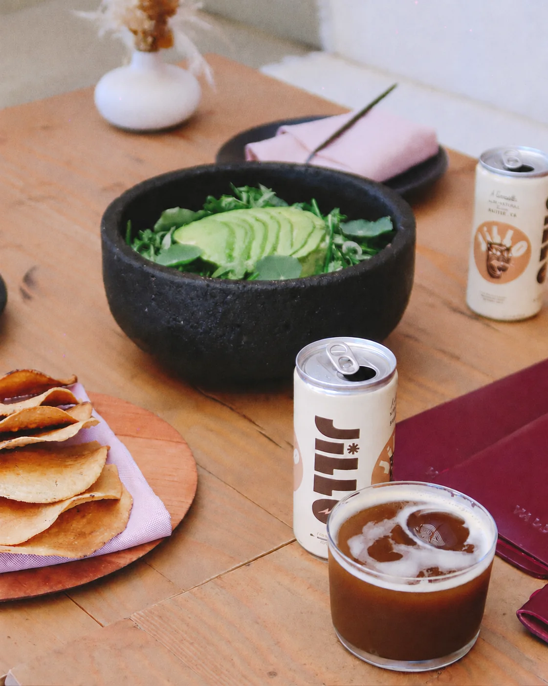 Two open JiLLO cans and an iced coffee cocktail beside avocado salad and tacos on a wooden table